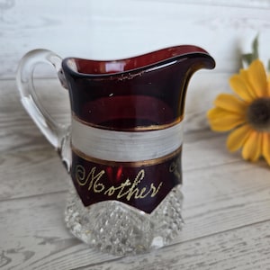 Puede incluir: Una jarra pequeña de vidrio transparente con asa, con borde rojo oscuro y una banda blanca. La palabra "Mother" está escrita en letras doradas en la parte delantera. La base tiene un patrón de corte de diamante. Una flor amarilla está en el fondo.