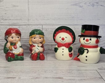 Op de afbeelding: Vier keramische beeldjes waaronder een kind met een rode muts en hobbelpaard, een kind met een groene muts en pop, een sneeuwpop met een rode muts en strik, en een sneeuwpop met een zwarte hoge hoed met rode band en hulst.