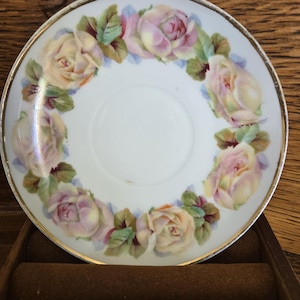 May include: A white porcelain saucer with a hand-painted floral design. The design features a ring of pink and yellow roses with green leaves around the rim, and a gold trim. The saucer is set on a brown surface.