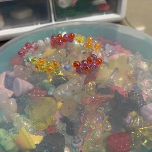 Puede incluir: Un cuenco de plástico transparente lleno de una variedad de cuentas y abalorios de colores. Una pulsera de cuentas de colores del arcoíris se encuentra encima del surtido. Las cuentas son de varias formas y tamaños, incluyendo estrellas, flores y corazones. Los colores incluyen rosa, naranja, amarillo, verde y morado.