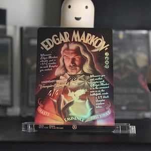 Puede incluir: Una carta coleccionable con Edgar Markov, un Caballero Vampiro. La carta tiene un retrato de un hombre con pelo largo y blanco y texto que incluye "Prisa", "Eminencia" y "Primer golpe". La carta se muestra en un soporte acrílico transparente.