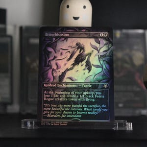 May include: A holographic "Bitterblossom" trading card, featuring a faerie illustration with text. The card is displayed in a clear acrylic stand. The card's text includes the phrase "Kindred Enchantment - Faerie".