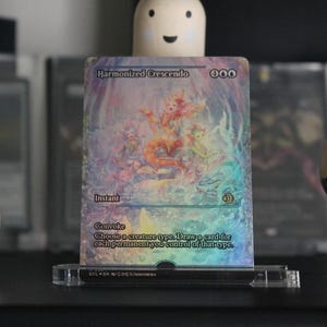 Könnte beinhalten: Eine holografische Sammelkarte mit dem Titel "Harmonized Crescendo" wird in einem transparenten Acrylständer präsentiert. Die Karte zeigt farbenfrohe Fantasy-Kunst mit dem Text "Instant" und "Convoke".