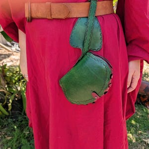 May include: A green leather pouch with a decorative leaf-shaped flap, attached to a brown leather belt. The pouch is worn over a red garment. The pouch has a circular shape with red accents.