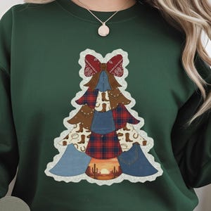 Könnte beinhalten: Forstgrünes Sweatshirt mit einem Weihnachtsbaum-Design. Der Baum besteht aus Jeans-, Karo- und Cowboy-Stiefel-Elementen, gekrönt von einer roten Schleife. Die Basis des Baumes zeigt eine Wüsten-Sonnenuntergangsszene. Die Worte "Forest green" sind auf einem herzförmigen Etikett aufgedruckt.