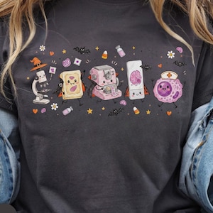 Könnte beinhalten: Ein dunkelgraues T-Shirt mit einem Grafikdruck von Cartoon-Laborgeräten und Zellen in Halloween-Kostümen, darunter ein Mikroskop mit Hexenhut, ein Toaster-Gebäck, eine rosa Maschine, ein Objektträger mit Gehirn und eine Zelle.