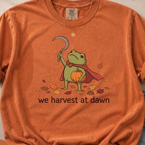 Könnte beinhalten: Ein orangefarbenes T-Shirt mit kurzen Ärmeln und einer Grafik eines grünen Frosches, der einen roten Umhang trägt, eine Sichel hält und einen orangefarbenen Kürbis trägt. Das Design enthält Herbstblätter und den Text we harvest at dawn.