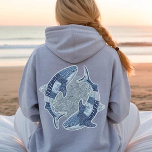 May include: A pale grey hoodie with a design of two blue whale sharks on the back. The whale sharks have a patchwork design with white polka dots. The hoodie has a drawstring hood and long sleeves.