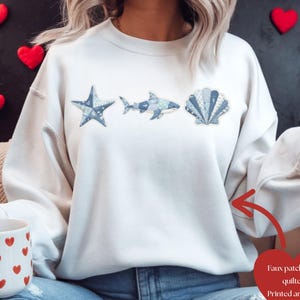Könnte beinhalten: Weißes Sweatshirt mit einem Patchwork-Design in Blau, das einen Seestern, einen Hai und eine Muschel zeigt. Das Sweatshirt hat einen Rundhalsausschnitt und lange Ärmel. Das Kunstwerk ist auf der Vorderseite aufgedruckt. Der Text auf dem Bild lautet "Faux patchwork quilted Printed artwork".
