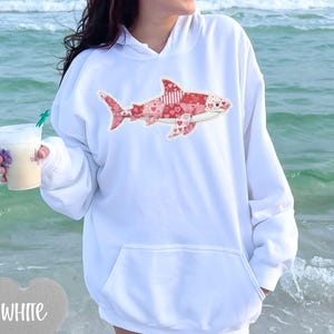 Op de afbeelding: Witte hoodie met een haaienontwerp in rood- en roze tinten, met hart- en streepmotieven. De hoodie heeft een voorzak en een capuchon met trekkoord. Het woord "WHITE" staat in een hartvorm linksonder.
