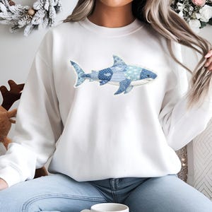 May include: White sweatshirt with a blue shark design. The shark is made up of different blue patterns, such as polka dots and stripes. The sweatshirt is a casual item of clothing, suitable for everyday wear.