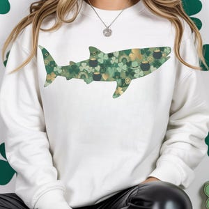 Puede incluir: Una sudadera blanca de cuello redondo con un gráfico de silueta de tiburón en el pecho. El tiburón está relleno con un patrón repetitivo de tréboles verdes, monedas de oro, calderos negros y pequeños arcoíris sobre un fondo verde claro.