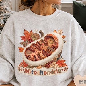 May include: A light gray crewneck sweatshirt featuring a graphic of a mitochondrion surrounded by autumn leaves. The mitochondrion is filled with orange and brown patterns, labeled with the text ATP and POWERHOUSE. The word Mitochondria is printed below.