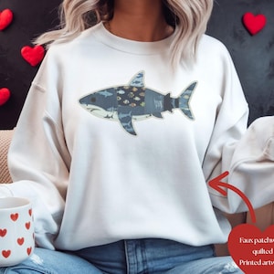 May include: A white sweatshirt featuring a faux patchwork shark design. The shark is primarily blue with various fish and aquatic patterns. The text "Faux patchwork quilted Printed artwork" is also visible.