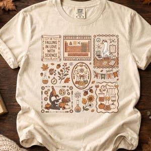 May include: A cream-colored t-shirt featuring a graphic print with the text 'FALLING IN LOVE WITH SCIENCE'. The design includes a periodic table, a goose in a lab coat, a microscope, various laboratory glassware, autumn leaves, and pumpkins.