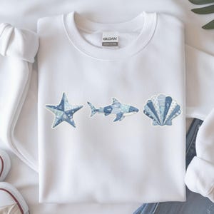 May include: A white crewneck sweatshirt featuring a central graphic print of a starfish, a shark, and a seashell. Each marine creature is designed with a patchwork pattern in various shades of blue and white, including stripes and dots.