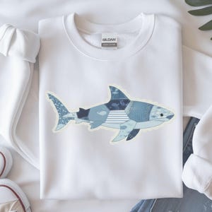 Op de afbeelding: Wit sweatshirt met een haaienontwerp. De haai is samengesteld uit verschillende blauwe denim patches, waaronder strepen en vispatronen. Het sweatshirt heeft een ronde hals en lange mouwen, perfect voor casual wear.