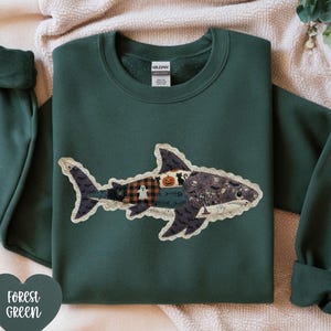 May include: A forest green crewneck sweatshirt featuring a large shark-shaped applique on the chest. The shark is constructed from various fabric patches with Halloween-themed patterns, including pumpkins, ghosts, bats, and spiderwebs.