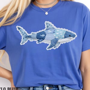 May include: Blue t-shirt with a shark design. The shark is made of various blue patterned fabrics, including stripes, polka dots, and floral prints, with a white outline. The text "FLO BLUE" is at the bottom.