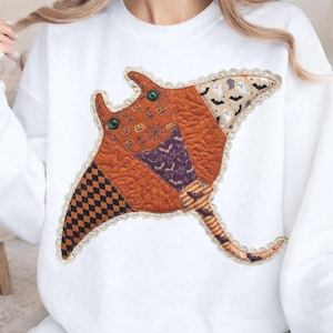 Könnte beinhalten: Ein weißes Sweatshirt mit Rundhalsausschnitt und einer großen, gesteppten Applikation in Form eines Rochen. Der Rochen besteht aus verschiedenen Stoffstücken mit orangefarbenen Kürbis-, lila Fledermaus- und beigen Geistermustern.