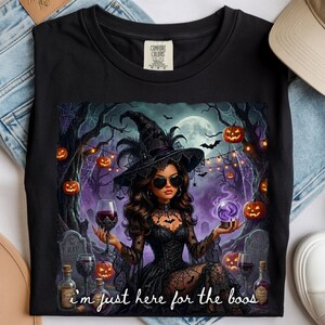 May include: A black t-shirt featuring a graphic of a witch wearing a hat and sunglasses, holding a wine glass and a crystal ball. The scene includes pumpkins, bats, trees, and tombstones with the text "I'm just here for the boos" at the bottom.