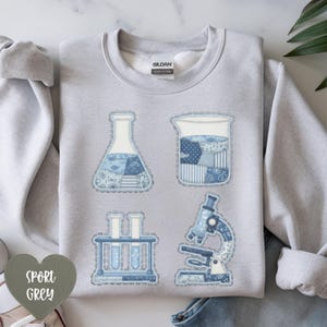 May include: Sport grey sweatshirt featuring four blue science-themed patches: a flask, beaker, test tubes, and a microscope. The patches have a patchwork design with denim-like textures and decorative stitching. The sweatshirt has a crew neck and long sleeves.