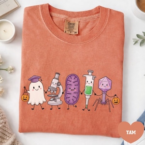 May include: A terracotta-colored short-sleeve t-shirt featuring a graphic of five cartoon characters holding hands: a ghost in a graduation cap with a pumpkin bucket, a microscope, a purple mitochondrion, a syringe, and a bacteriophage with a pumpkin.