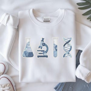 Puede incluir: Sudadera blanca con gráficos de temática científica. El diseño incluye un vaso de precipitados, un microscopio, un tubo de ensayo y una cadena de ADN, todos en tonos azules. La sudadera está hecha de un material suave y es adecuada para uso casual.