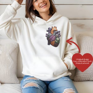 May include: A cream-colored hooded sweatshirt featuring a graphic of an anatomical heart on the chest. The heart design incorporates various patterns and textures. A red heart-shaped graphic includes the text: Original design, Faux embroidery look, Printed artwork.