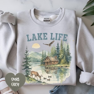 May include: A light gray crewneck sweatshirt featuring the text LAKE LIFE in blue letters. The graphic depicts a watercolor-style landscape with a wooden cabin, a deer, ducks on a lake, pine trees, mountains, and a flying eagle in the sky.