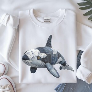 May include: White sweatshirt with a large orca whale design. The whale is made of denim patches and nautical-themed fabric, featuring a lighthouse and compass rose. The sweatshirt has a crew neck and long sleeves.