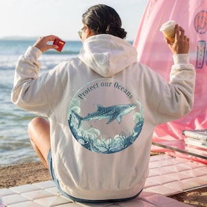 Könnte beinhalten: Cremefarbener Kapuzenpullover mit einem kreisförmigen Aufdruck auf dem Rücken, der einen Walhai, Meereswellen und den Text "Protect our Oceans" zeigt. Der Kapuzenpullover hat eine Kapuze und lange Ärmel. Die Person im Bild hält ein kleines rotes Objekt und eine Tasse.