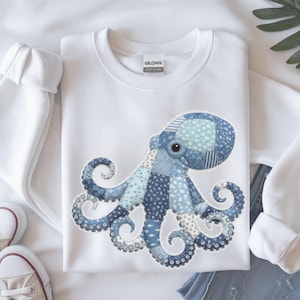 May include: White sweatshirt featuring a patchwork octopus design in shades of blue. The octopus has detailed tentacles and a large eye. The sweatshirt is made of a soft material and is suitable for casual wear.
