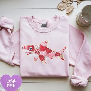 Puede incluir: Una sudadera de manga larga de color rosa claro con un gráfico central de un tiburón compuesto por varios parches con patrones de corazones rojos, blancos y rosas. El diseño del tiburón incluye el texto XOXO en la cola y cuello redondo.