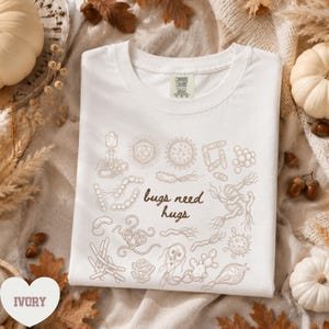 Könnte beinhalten: Ein elfenbeinfarbenes T-Shirt mit einer braunen Strichgrafik verschiedener Mikroorganismen und Bakterien. Der Text "bugs need hugs" ist mittig in einer Schreibschrift gedruckt. Das T-Shirt hat einen Rundhalsausschnitt und ein Comfort Colors Etikett.