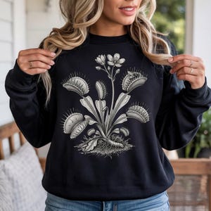 May include: A black long-sleeve sweatshirt featuring a detailed white graphic print of a Venus flytrap plant with multiple open traps, leaves, and a central flower stalk, centered on the front of the garment with a crew neckline.