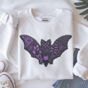 May include: A white long-sleeve sweatshirt featuring a large bat-shaped graphic on the chest. The bat design is composed of various purple and black patterned sections, including ghosts, skulls, spiderwebs, bats, and a cat face, with a black lace trim.