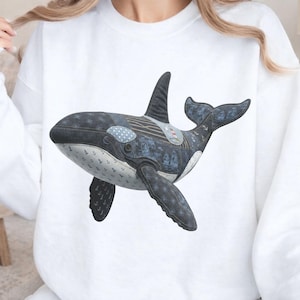 May include: White sweatshirt featuring a detailed illustration of a killer whale. The orca design incorporates various denim patterns, including polka dots and nautical motifs. The whale's body is primarily dark blue and gray, with white accents.
