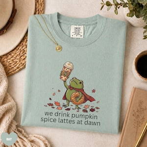 Op de afbeelding: Een licht saliegroen t-shirt met een afbeelding van een groene kikker in een rode cape, die een pumpkin spice latte en een donut vasthoudt. De tekst luidt, we drink pumpkin spice lattes at dawn. Comfort Colors label in de nek.