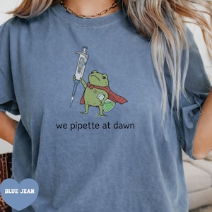 Peut inclure: Un t-shirt bleu jean avec une illustration d'une grenouille verte portant une cape rouge, tenant une pipette et un bécher contenant un liquide vert. Le texte sous la grenouille indique we pipette at dawn en lettres minuscules noires.