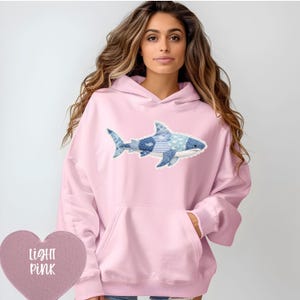 Puede incluir: Sudadera con capucha rosa claro con un diseño de tiburón azul. El diseño del tiburón incorpora varios patrones azules, incluyendo rayas y lunares. La sudadera tiene un bolsillo de canguro y una capucha con cordón.