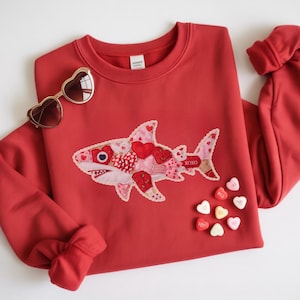 May include: Red sweatshirt featuring a shark applique made from hearts and fabric. The shark has a white lace trim and the word "XOXO" on its tail. Heart-shaped sunglasses and heart candies are also in the image.