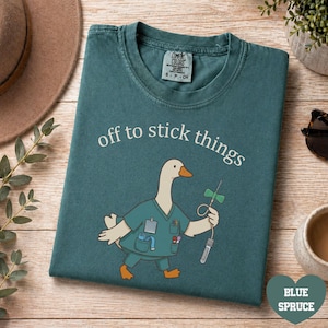 May include: A blue spruce t-shirt featuring a graphic of a goose dressed in medical scrubs holding a butterfly needle. The text above the goose reads "off to stick things" in white lettering. The shirt has a Comfort Colors brand neck label.