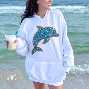 Puede incluir: Sudadera con capucha blanca con un gran diseño de delfín azul. El delfín está lleno de gemas azules y delineado en naranja. La sudadera tiene un bolsillo delantero y una capucha con cordón. La palabra "WHITE" está en forma de corazón.