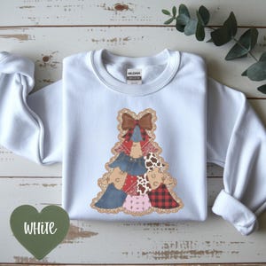 Puede incluir: Una sudadera blanca de manga larga con un gráfico de un árbol de Navidad formado por varios patrones, incluyendo mezclilla, cuadros rojos, estampado de vaca y pañuelos rojos, rematado con un lazo marrón y un borde de encaje beige.