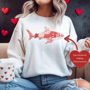 Op de afbeelding: Een witte sweatshirt met een haaienontwerp gemaakt van patchwork quilting in rood- en roze tinten. De haai is versierd met harten en andere patronen. De tekst "Faux Patchwork Quilting Printed artwork" is ook zichtbaar.
