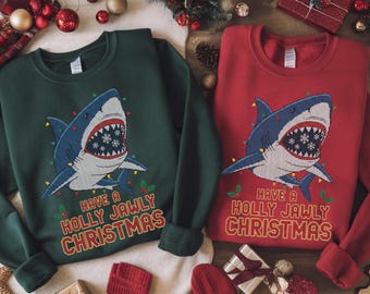 Puede incluir: Dos sudaderas, una verde y otra roja, presentan un diseño de tiburón con luces navideñas y el texto "Have a Holly Jawly Christmas". El tiburón tiene un cuerpo azul, dientes blancos y detalles de copos de nieve.