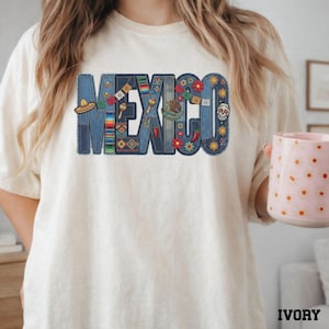 Puede incluir: Una camiseta color marfil con la palabra MEXICO en letras grandes con textura de mezclilla. Las letras están decoradas con ilustraciones coloridas que incluyen sombreros, maracas, chiles, flores, soles, una calavera de azúcar y banderines.