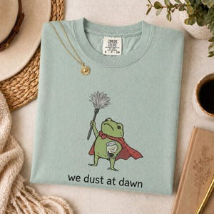 May include: A sage green t-shirt featuring a graphic of a green frog wearing a red cape, holding a feather duster and a jar labelled Powder Black. Below the frog, the text reads we dust at dawn. The shirt has a Comfort Colors brand neck label.