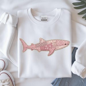 May include: White sweatshirt featuring a pink shark design. The shark is composed of various pink patterned fabrics, including leopard print and seashell motifs, outlined with a scalloped edge. The sweatshirt has a crew neck and long sleeves.